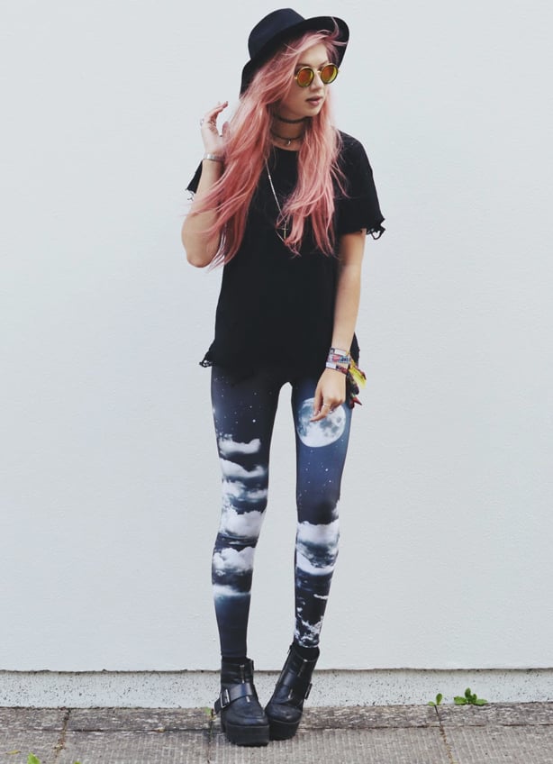 A young, pink-haired woman wearing night sky printed leggings, a black tee shirt and fedora hat