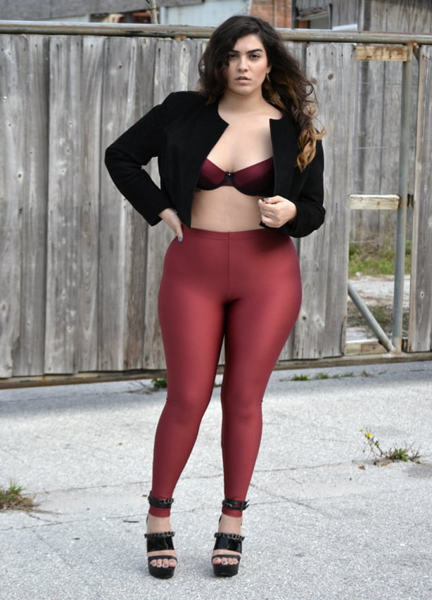 A plus-sized model wearing platform sandals, ruby red Lycra leggings, a black cropped jacket and red bra underneath 